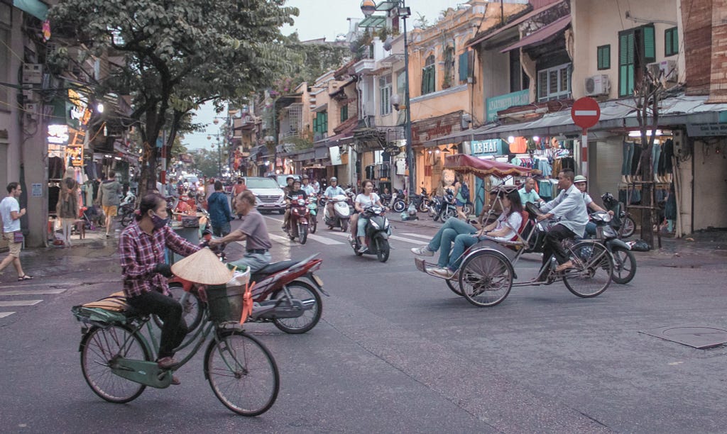 A photo of traffic in Hanoi, with various modes of transportation shown, including motorbikes, a bicycle, and a rickshaw