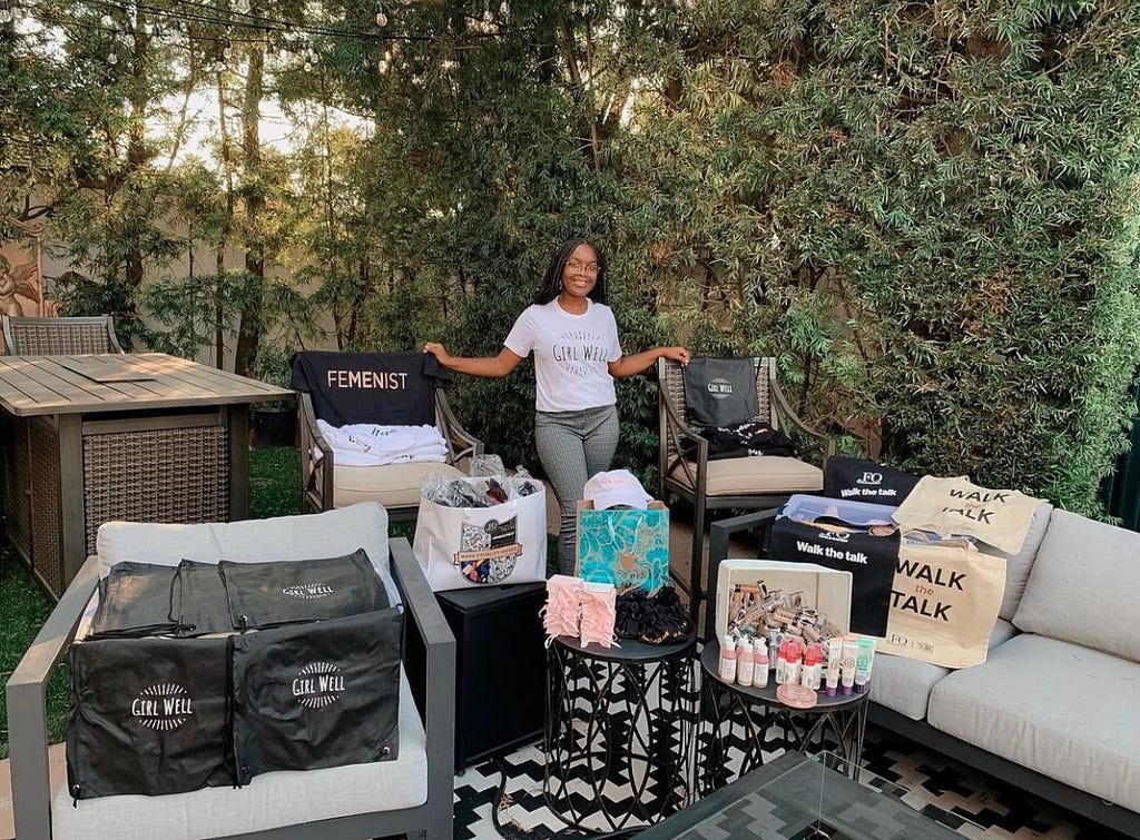 A young woman in a white “Girl Well” T-shirt poses in front of a table and chair area set up outdoors featuring boxes of self-care supplies. A blanket on one of the chairs reads “feminist.”