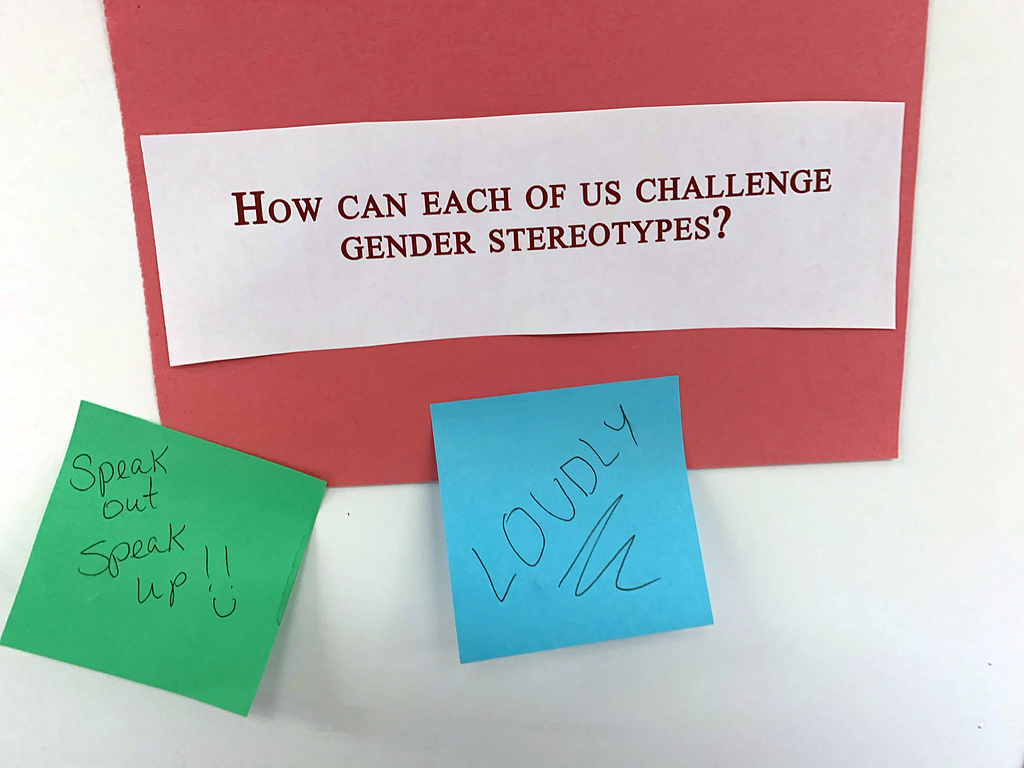 A sign reads, “How can each of us challenge gender stereotypes?” Two sticky notes answer “LOUDLY” and “Speak out, speak up!!”