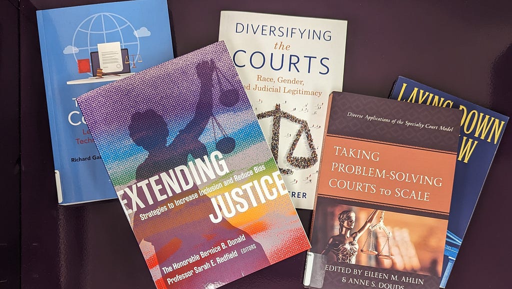 Five books are scattered over a dark purple background. One book has the title “Extending Justice” and the image on the cover is a silhouette of Lady Justice holding scales up with her left arm. There are multicolored stripes superimposed over the cover image. Another book has the title “Take Problem-Solving Courts to Scale.” It, too, has a photo of Lady Justice on the cover with books blurred in the background. The book title is printed in white text on a rust colored banner.