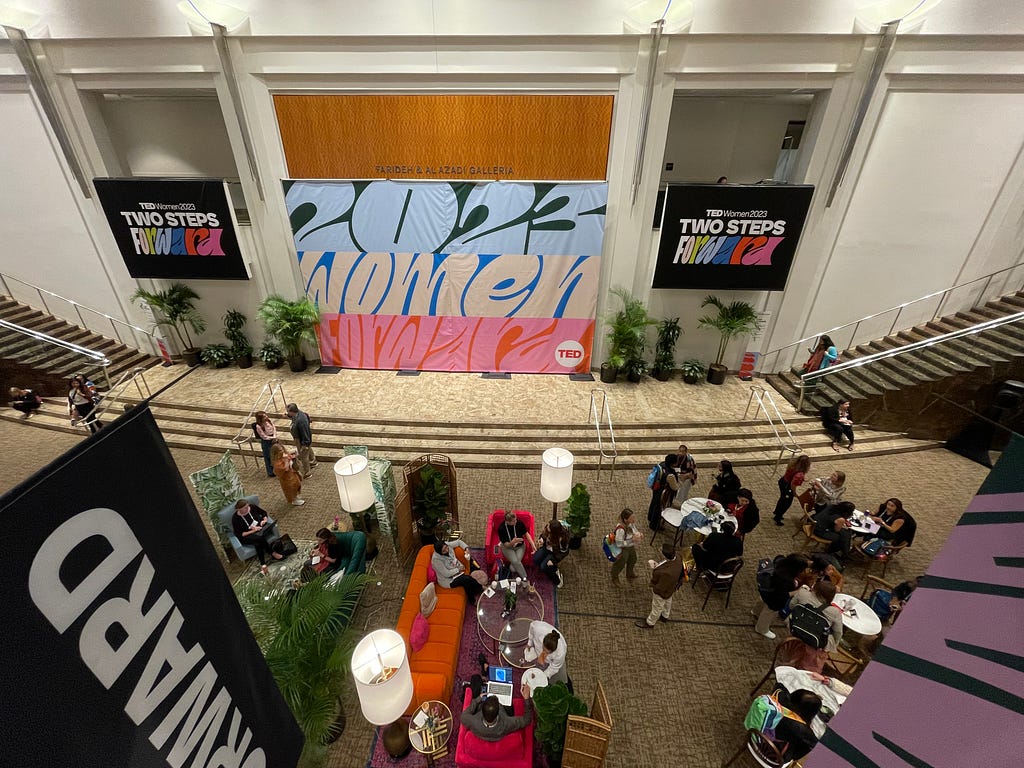 A photograph looking down at the TEDWomen2023 event space, with tall tables and lounges, plus signage for the event.