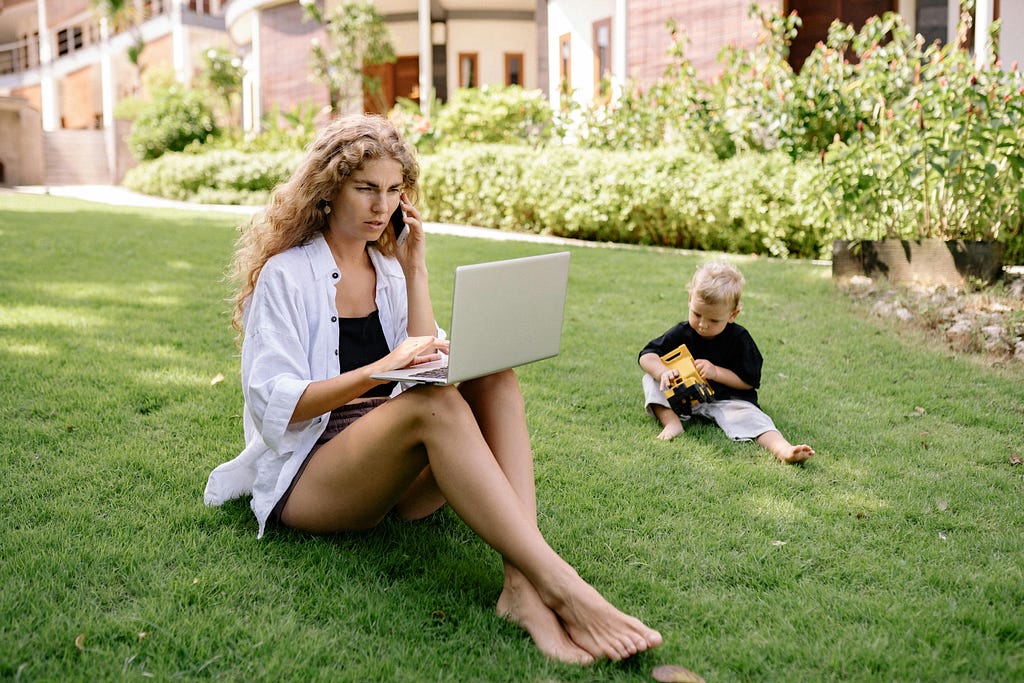 A mother working on a laptop, sitting on grass with her toddler, surrounded with lush plants and trees that cast ample amount of shadow.