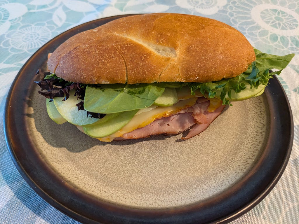 Ham, muenster, granny smith apple, and mixed salad greens on a hoagie roll, served on a beige plate with a brown rim. The tablecloth is light blue with a white floral pattern.