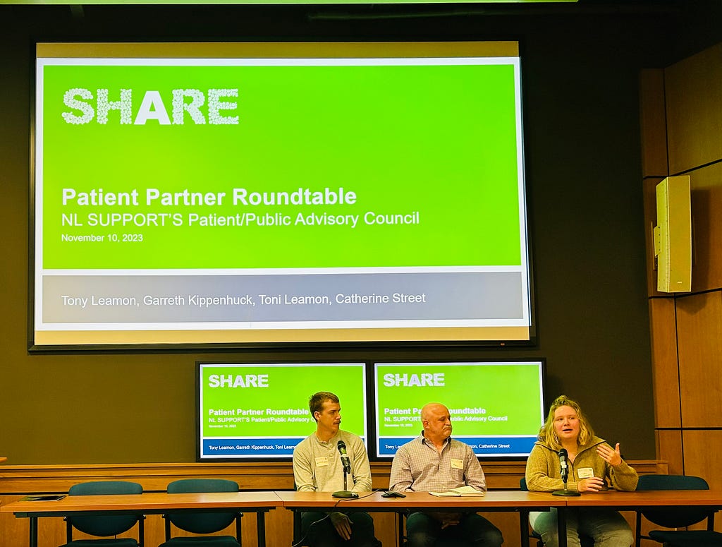 In a lecture hall, three patient partners from the NL SUPPORT Patient and Public Advisory Council (PPAC) participate in the Patient Partner Roundtable at SHARE Summit 2023. PPAC members are sitting at a table facing the audience and are answering questions from members of the audience. Behind the PPAC members are multiple screens displaying a PowerPoint slide.
