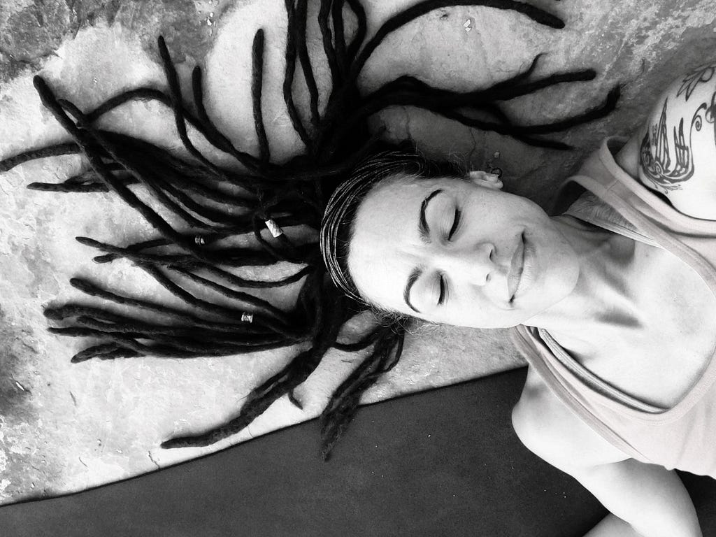 A smiling and serene woman with long dreadlocks lays on a yoga mat