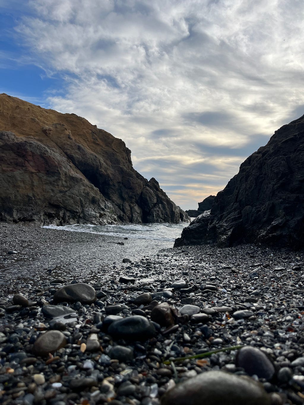 Water from the ocean advances between two rock formations in Mendocino, California. The perspective is low to the ground such that the small rocks on the beach appear enlarged.