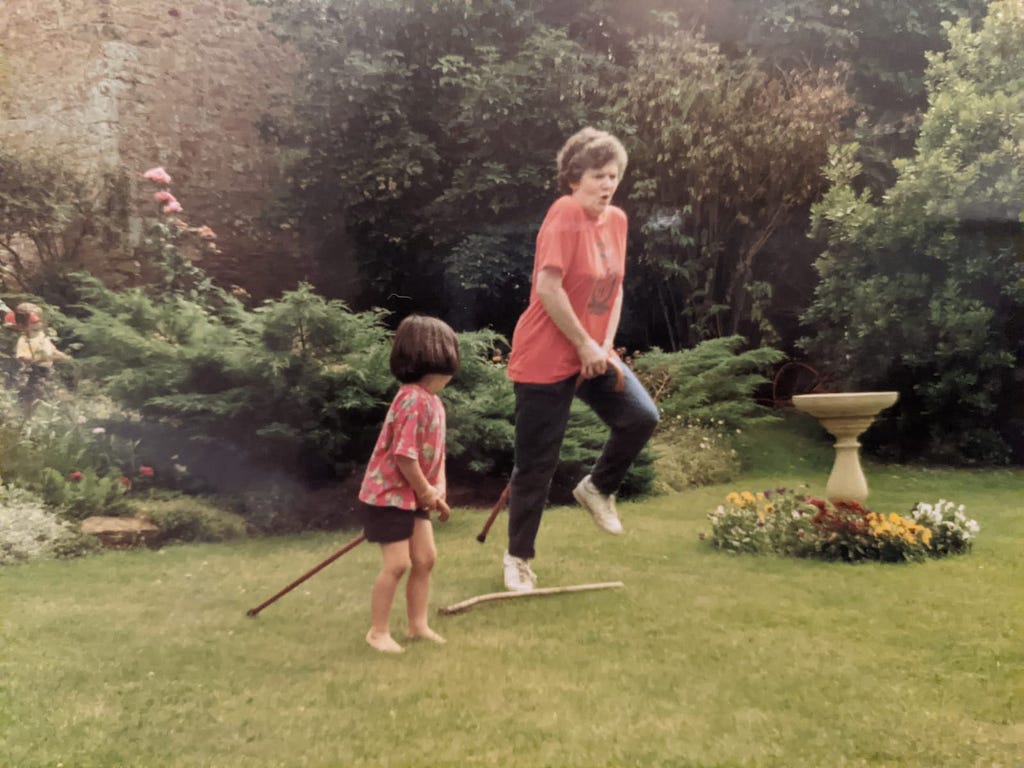 Young Sam playing make-belief horse-riding, with her grandmother in a garden.