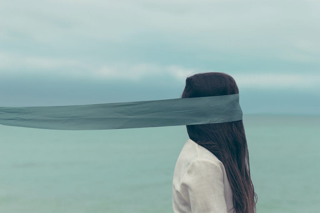 A woman’s face is obscured by a long, extended scarf that appears to be pulling her to the side. Her long black hair obscures the rest of her face. It is unclear which way she is facing.