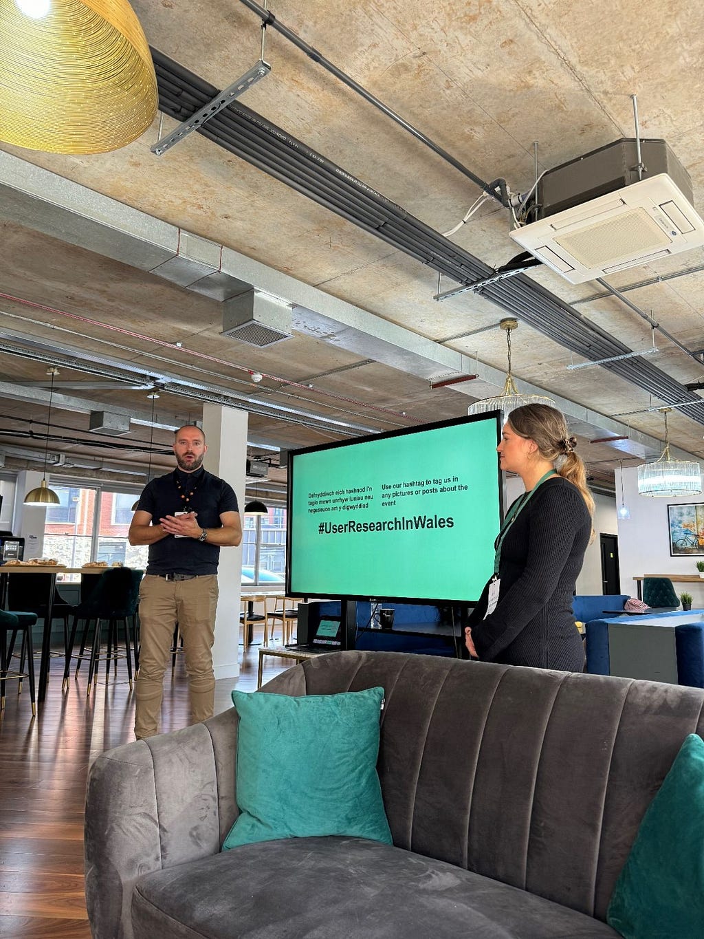 An office space with exposed ceiling and industrial lighting. Two people, a man and a woman, stand in front of a large screen displaying “#UserResearchInWales”. The man is gesturing while speaking. A gray couch with teal pillows is in the foreground. The space has a modern, open layout with wood flooring and stylish furniture.