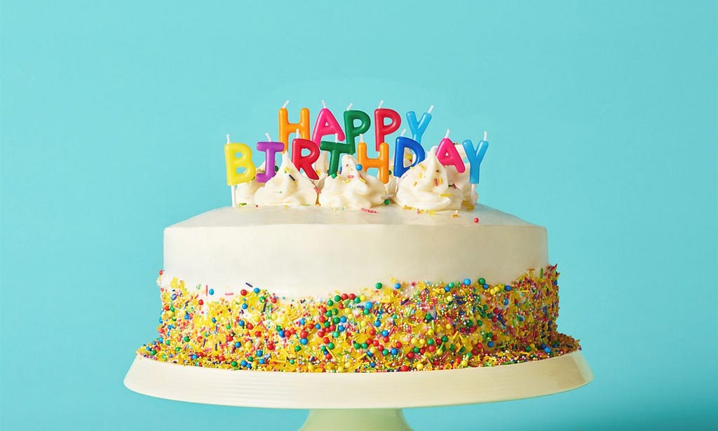 A turquoise blue background; in the foreground a white birthday cake with rainbow sprinkles around the base and multicoloured candles that spell happy birthday on the top.