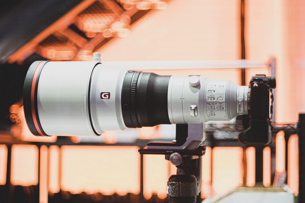 Modern, white-colored camera with a large, telephoto lens. The background of this image is a blurred orange.