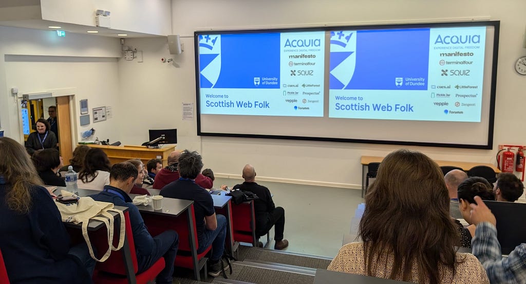 People in a lecture hall looking at screens with a slide welcoming them to the Scottish Web Folk conference