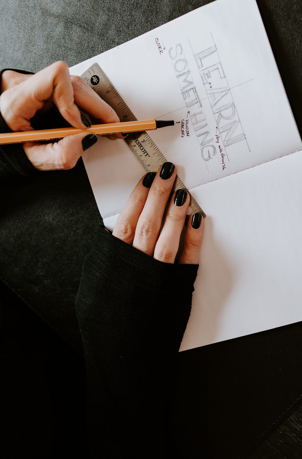 A designer uses a ruler to make visual design notes on a sketch. The sketch says “LEARN SOMETHING,” in uppercase block letters.