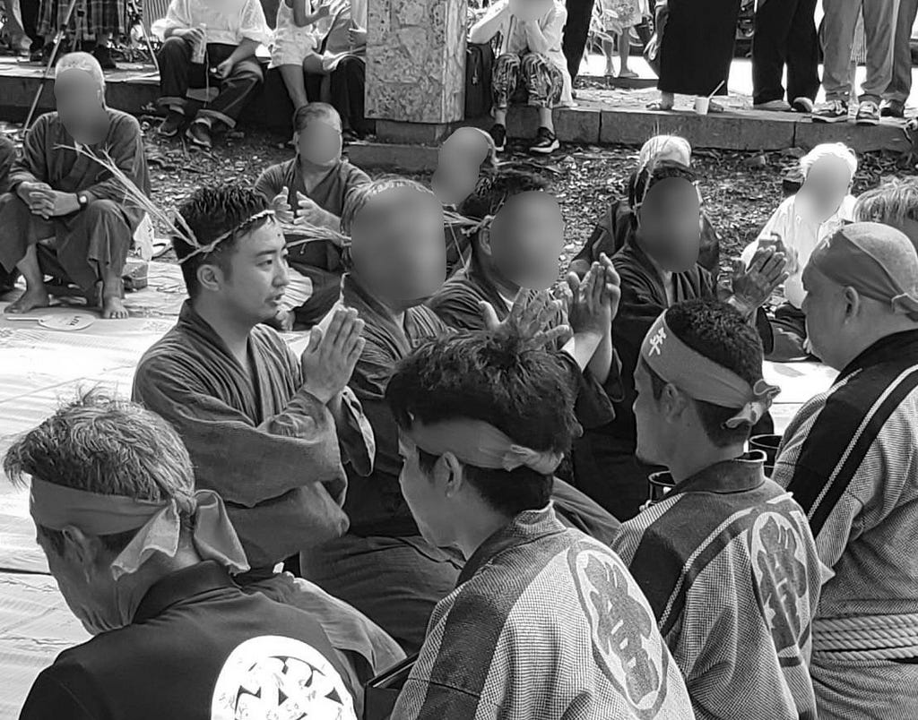 Participants at an outdoor ritual on Ishigaki Island, hands pressed together in prayer. In the center, a young man in dark blue traditional clothing wears a headband woven from grass — a sign of serving as a ritual officiant. Around him, men in dark kimono with white crest patterns and women in soft tones sit closely on tatami mats. Several wear red headbands marking their role. The air is quiet and concentrated; sunlight filters down through the trees above.