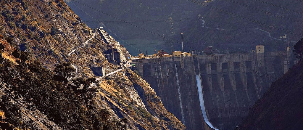 A steep-sided valley with small trees on the hillsides is shown, with sunshine on one slope, and Baglihar Dam mostly in shadow. A white stream of water flows through one of the dam’s sluice gates, and a small amount of the dam’s reservoir is visible in the background. Baglihar is a run-of-river power project on the Chenab River in the southern Doda district of the Indian state of Jammu and Kashmir. This is a closer crop of the photo included in the body text of the article.