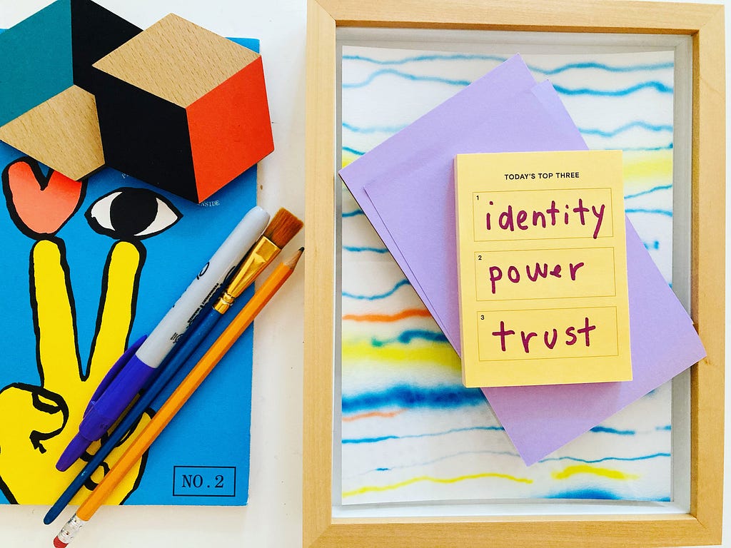 Tableau of a colorful magazine cover featuring an illustration of a hand making a peace sign, an eye, and a heart. A pencil, Sharpie, and paint brush lay on top of the magazine. A post-it with a hand-written list “ identity, power, trust” rest atop a framed piece of colorful line art.