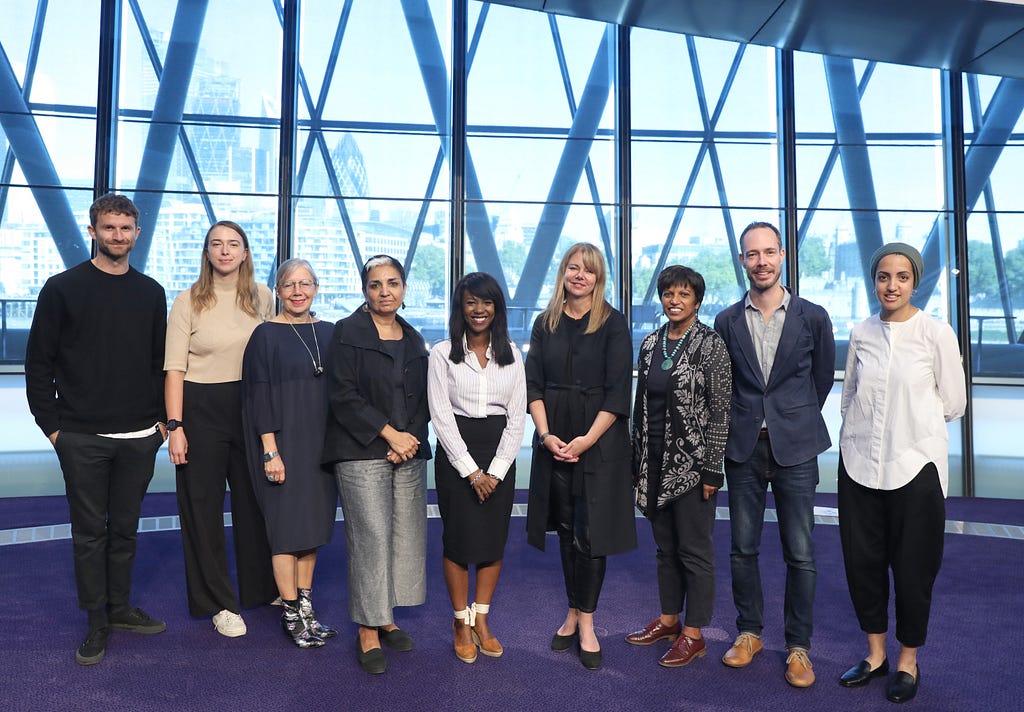 9 people stand on a purple carpet in front of a big window looking out onto the river Thames.