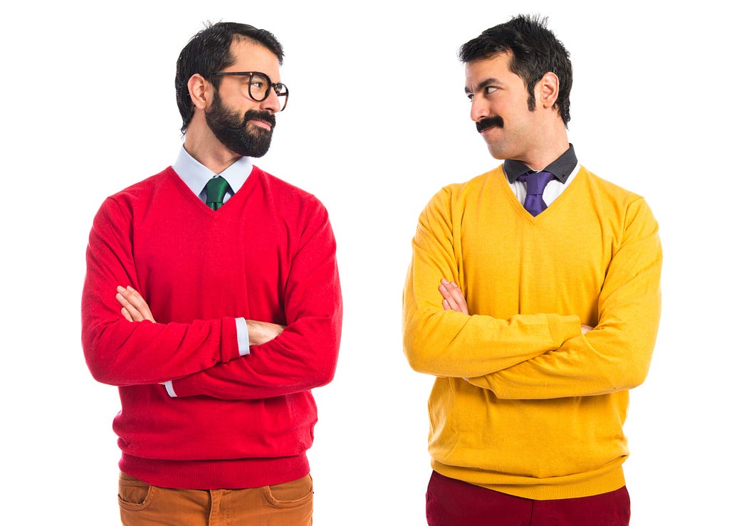 A imagem retrata dois irmãos gêmeos se encarando e sorrindo. O primeiro veste um suéter vermelho e o segundo, um amarelo.
