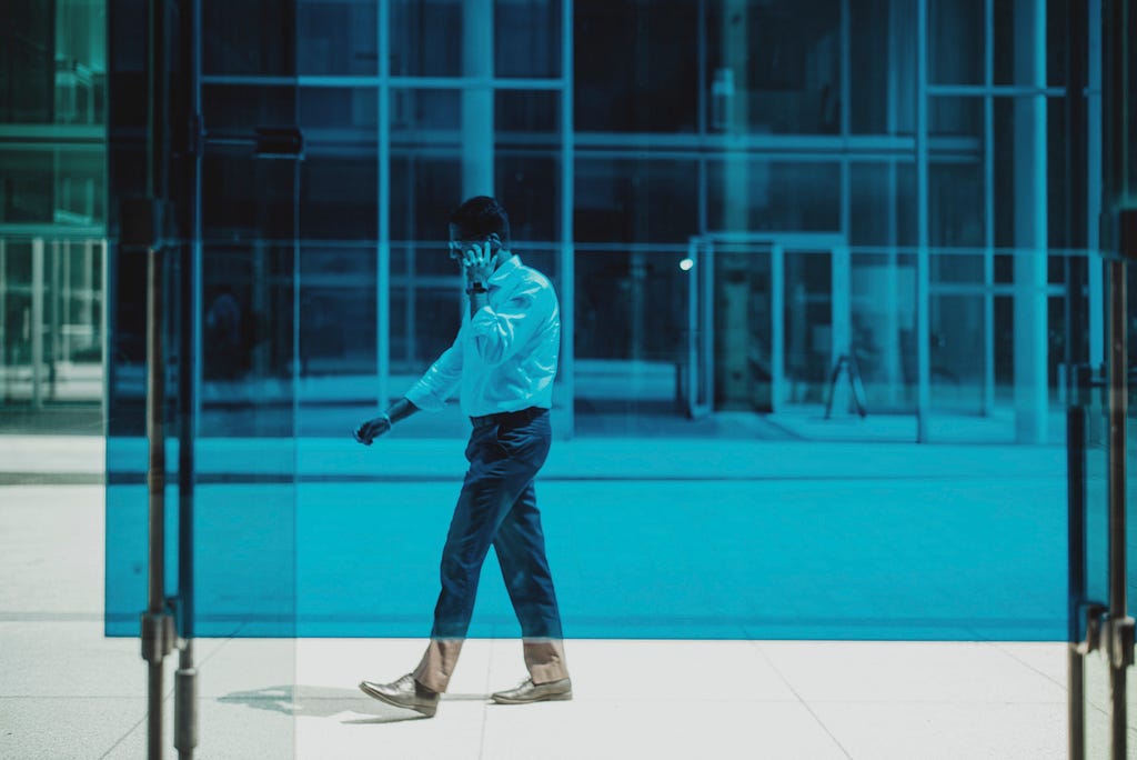 A man with a phone walking behind a blue glass.