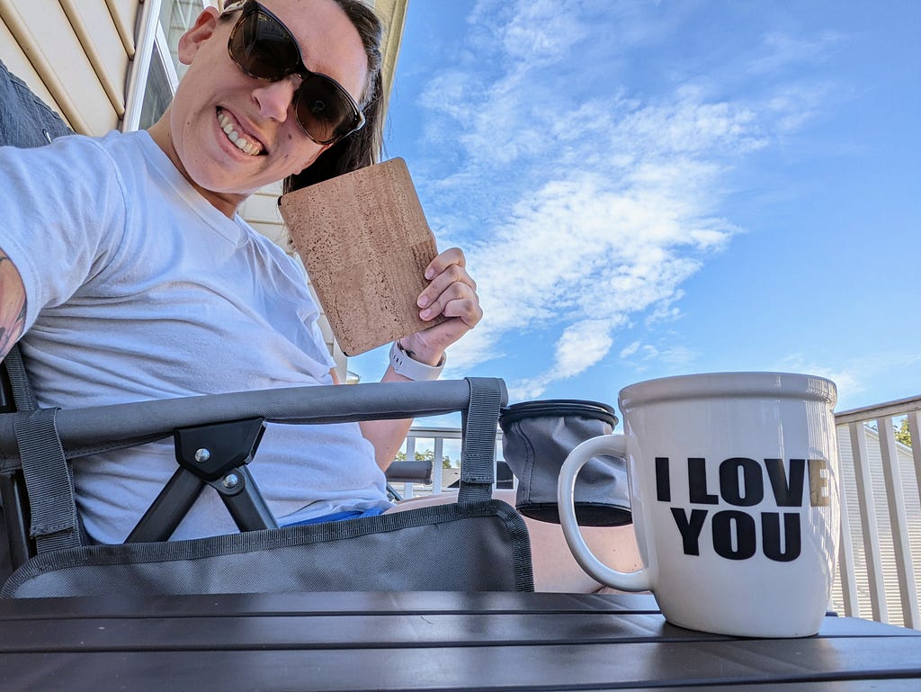 White female sitting outside in a gray camping chair. She is in a white t-shirt, has a large smile, and is holding up a Kindle which is in a cork case. She is wearing dark sunglasses and has a mug sitting near her that reads “I love you.”