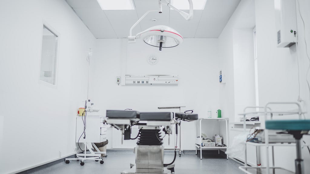 A photo of a surgical room. A dark bed is in the middle, on a hydraulic lift. A large lamp is positioned over the bed. A variety of tables and carts with supplies sit around the bed. The walls are white and the floor is gray.