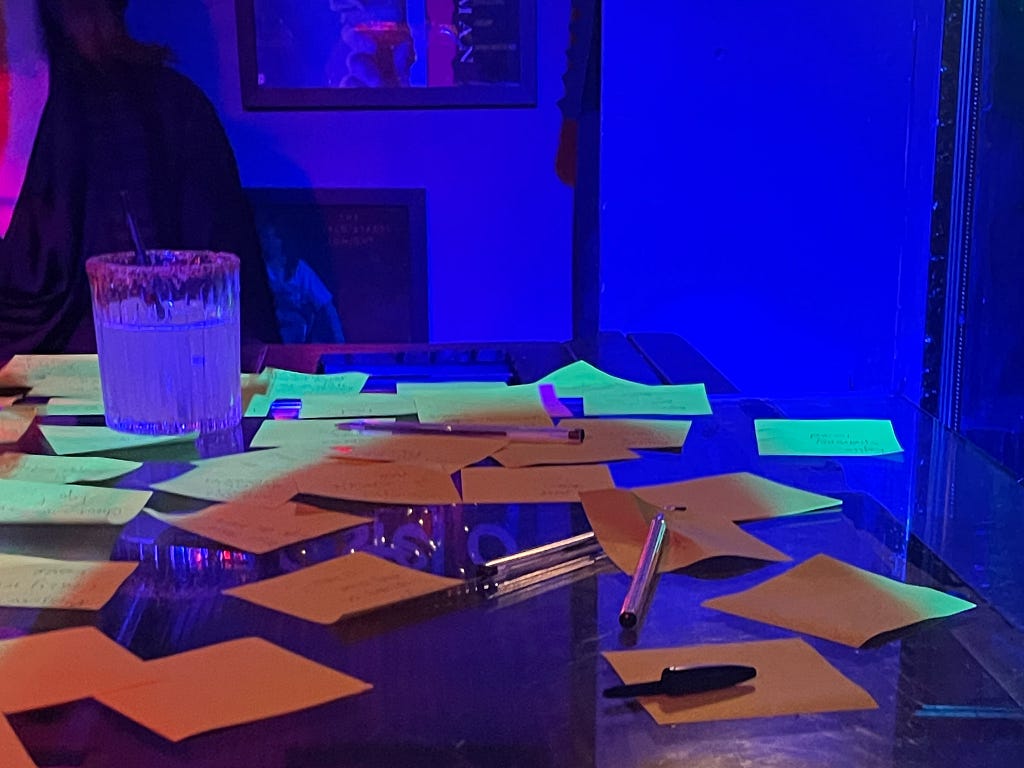 A dark bar, with lots of yellow post it notes and a pen laid out on the top of a piano, alongside a drink