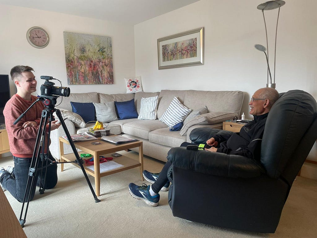 Mohamed, an older man, sits in an armchair in his lounge. He laughs as a younger man sits opposite filming him.