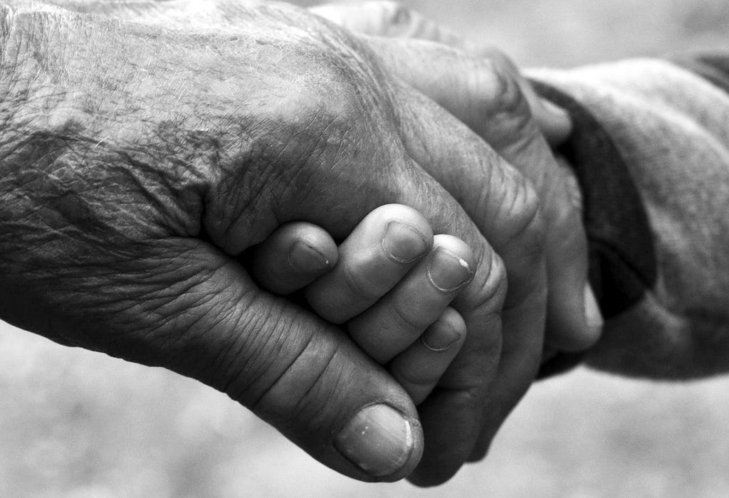 Black and white picture depicting two hands holding to each other. One is old (on the left) and one is that of a child (on the right).