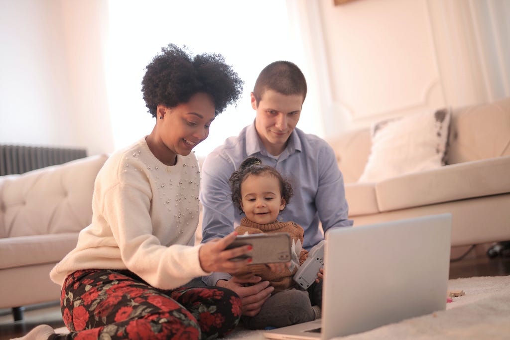 Family of three watching something on a phone with a laptop in front of them.