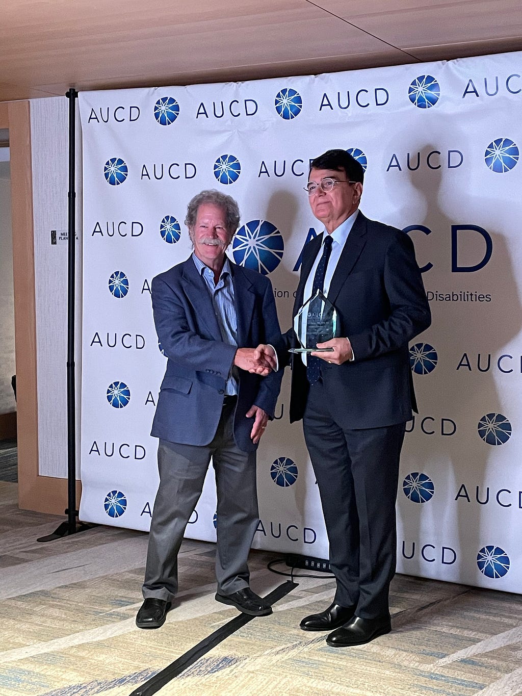 Two white men shaking hands in front of a screen displaying the words “AUCD”; one of the men is holding an award.