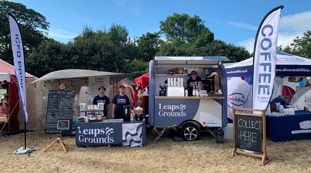A festival coffee stall from Leaps & Grounds, with a branded table and coffee cart, a white flag saying ‘coffee’, and two blackboard signs (a menu and a ‘collect here’ sign). Four people stand ready to serve coffee.