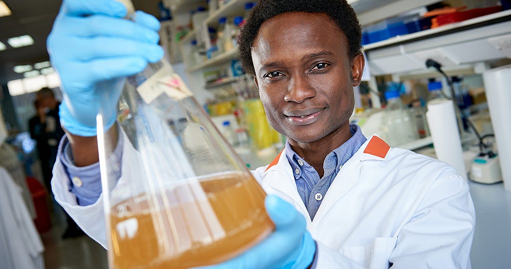 Dr Oluwapelumi Adeyemi wearing a white lab coat and blue gloves holding a beaker with liquid in it