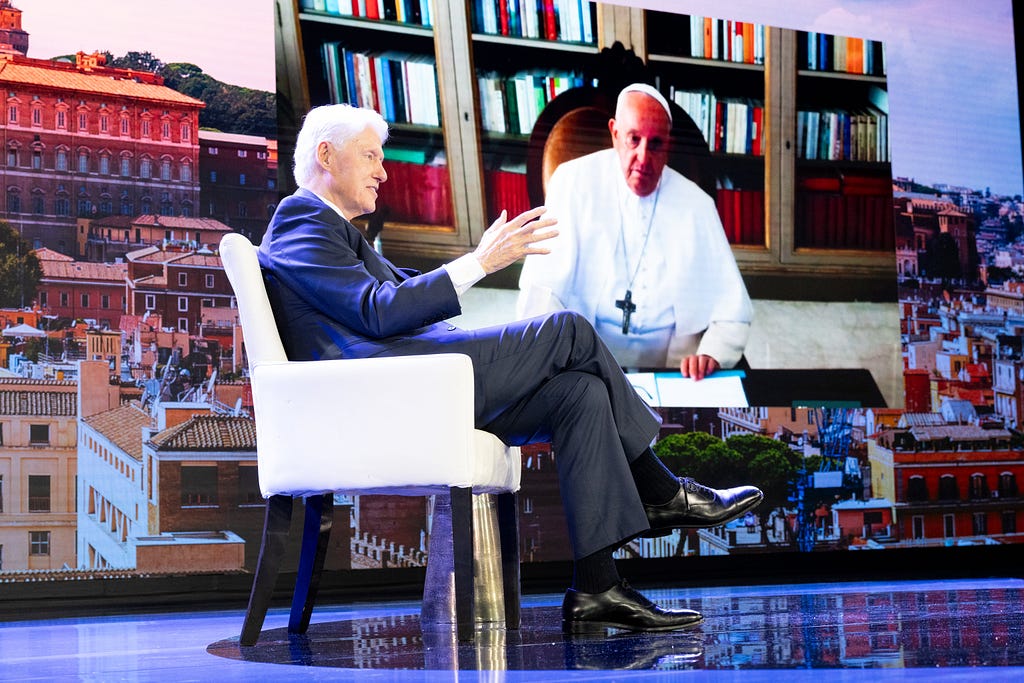 President Clinton sits in a white chair speaking to Pope Francis who is on a large screen behind him.