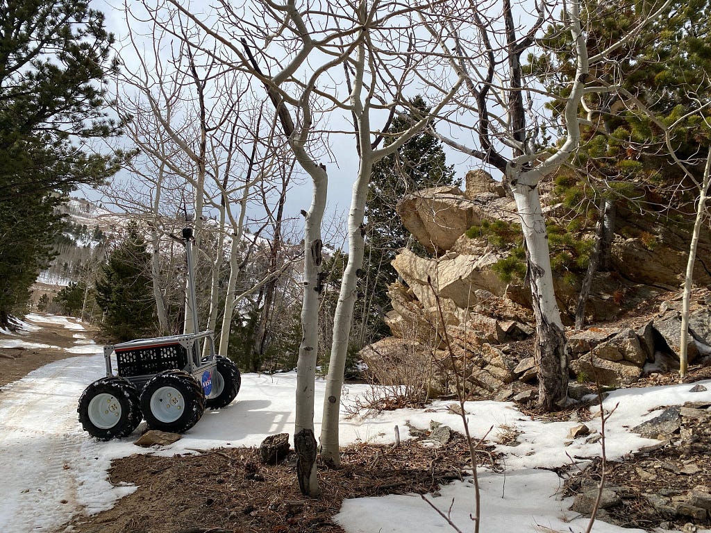A square robot with four rugged wheels and a tall mask holding a camera in front of an exposed rock outcrop. The rover is approximately the size of the trunk of an SUV. The landscape is mostly flat with a thin layer of snow.
