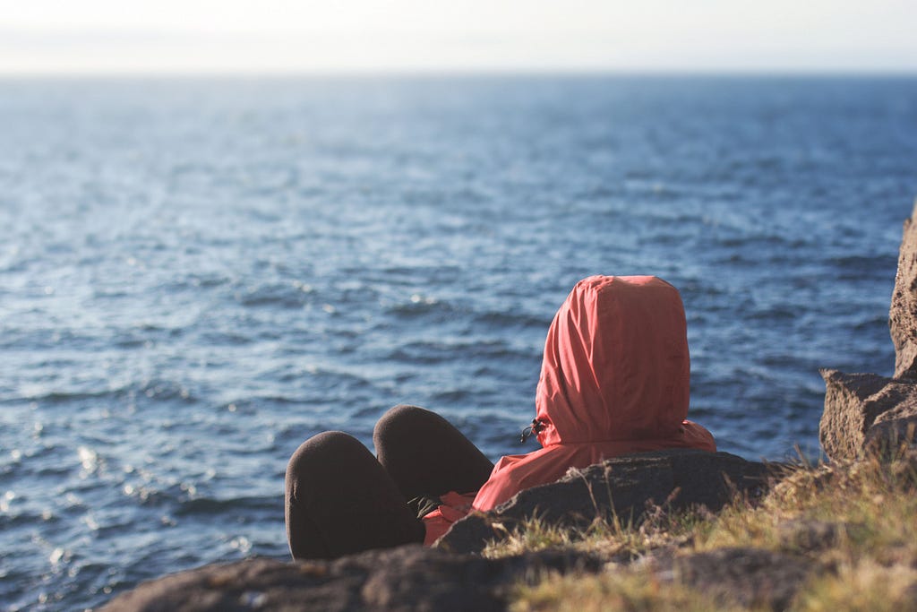 Photo of a person wearing a hoodie leaning on some rocks at the beach and looking out at the ocean