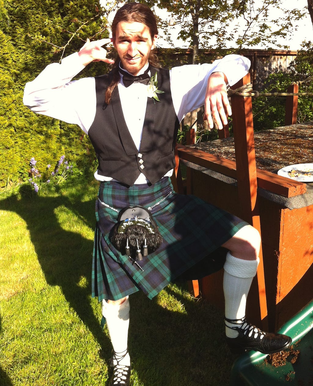 Me in a blue/green/black kilt, with white shirt, black vest, sporran pouch, thick white socks, and open black dress shoes. I’m making a ‘call me’ gesture with one hand, while leaning and stepping on a structure.