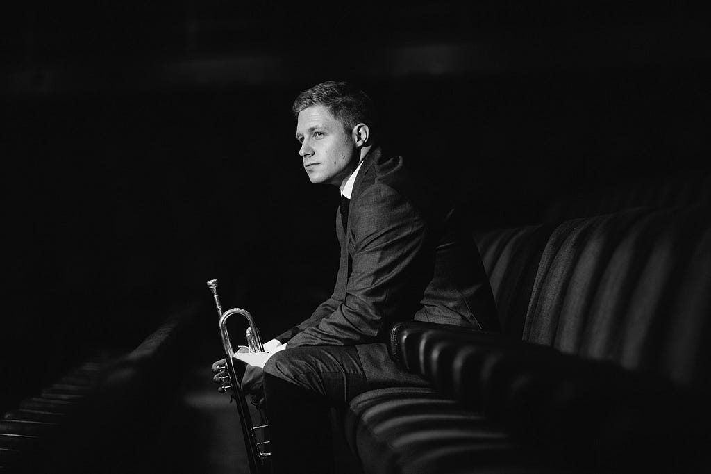 Black and white photo of Philip Cobb in a suit sitting in a row of seats, holding a trumpet