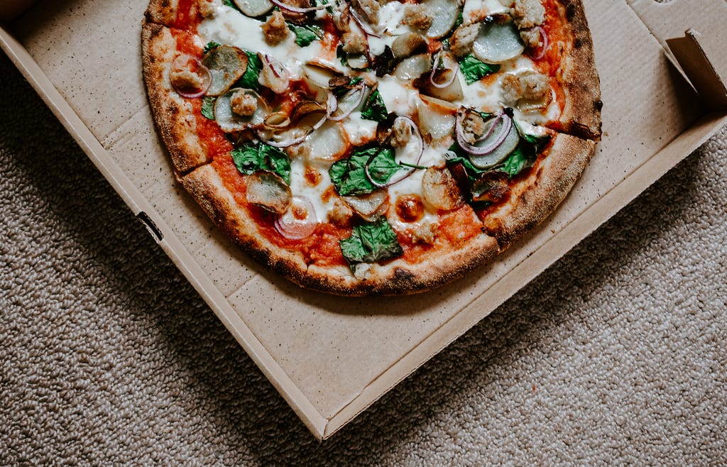 A pizza with various toppings of onions, basil, cheese and more sits in a carboard box. The box sits on a fuzzy light brown surface.