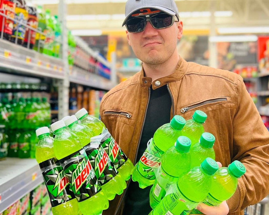 Man in a baseball cap and sunglasses standing in Walmart and holding two 6 packs of Mountain Dew
