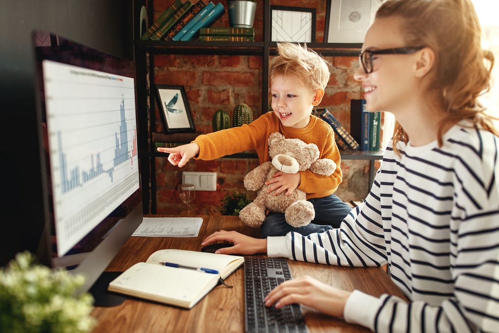 A woman and a toddler are at a desk looking at a computer sceen. She is smiling as he points at the screen.