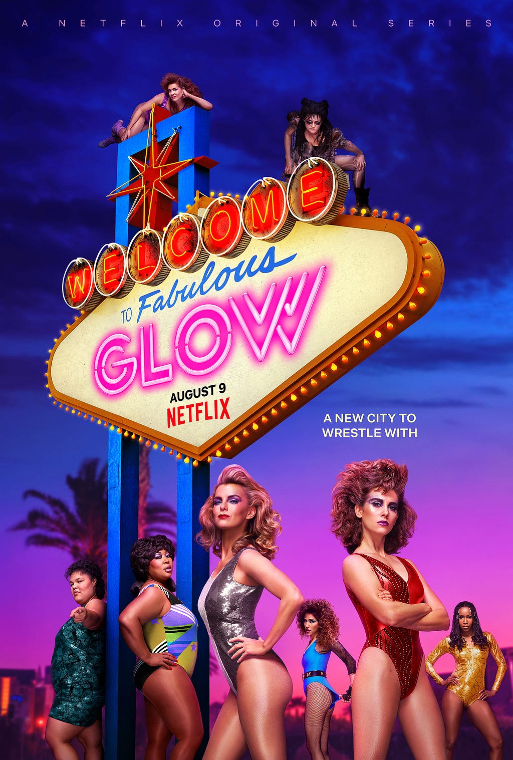 The cast of GLOW dressed in their 80s wrestling outfits around a large illuminated scene like the sort you might find in a classic American motel. Several are stood in front of the sign, others are actually stood on the sign itself