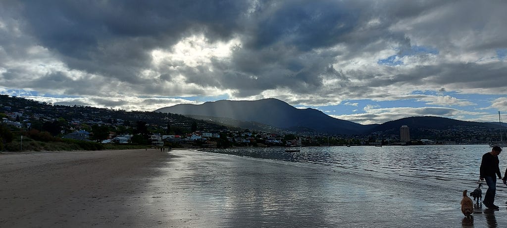 view of Hobart city from Long Beach, Sandy Bay. it is almost night, the sun is setting, but the sky is cloudy, with sun coming through in patches. The water is calm and glassy. The hills are dark except for one in the middleground, where the author’s home is situated.