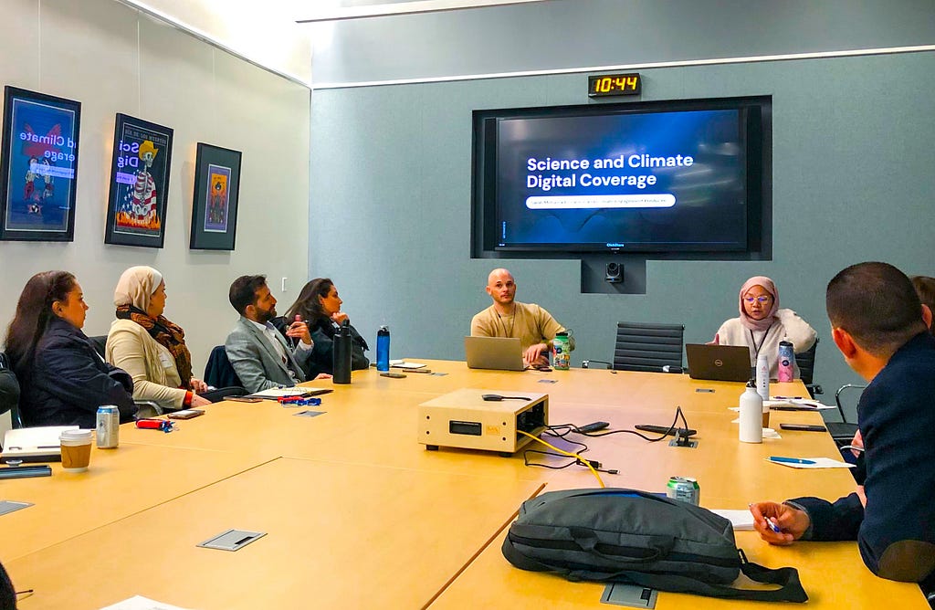 Adults sitting at a long rectangular conference table listening to a presentation on a wall monitor titled “Science and Climate Digital Coverage.”