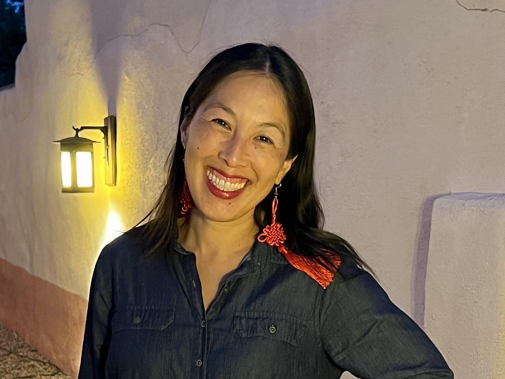 Photograph of the author standing in front of a white wall. She has straight black hair and long red tassel earrings. She is smiling.