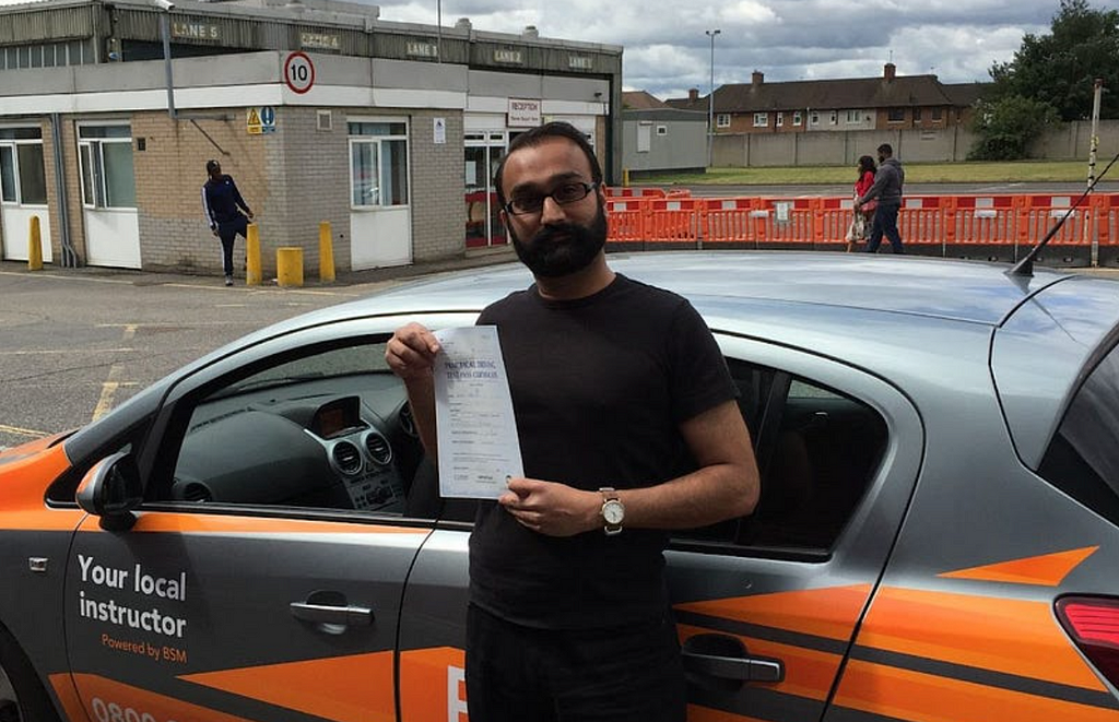 Brown man with beard and glasses in black clothes standing in front of a a learner car holding a piece of paper.