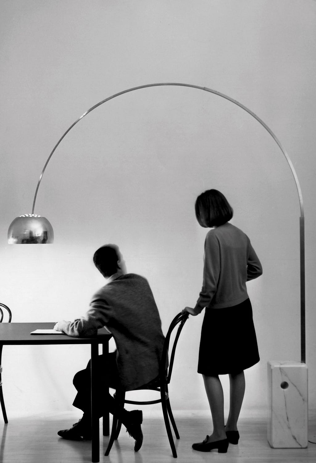The Arco lamp designed for the studio Flos positioned above a dining table where a man is sitting and a woman standing next to him. The lamp creates an arc above them and shines light onto the table.