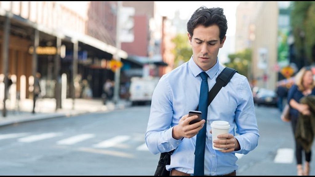 Half-portrait of businessman walking down street wearing blue button down shirt and tie, with messenger bag draped on shoulder, frowning at cell phone in one hand and carrying take-out coffee in the other hand.