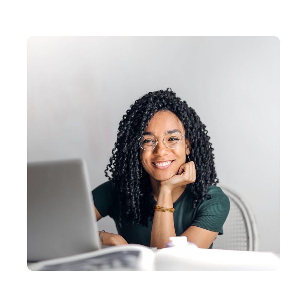 Picture of a woman in a green shirt with curly hair smiling while sitting at a desk with a laptop on it.