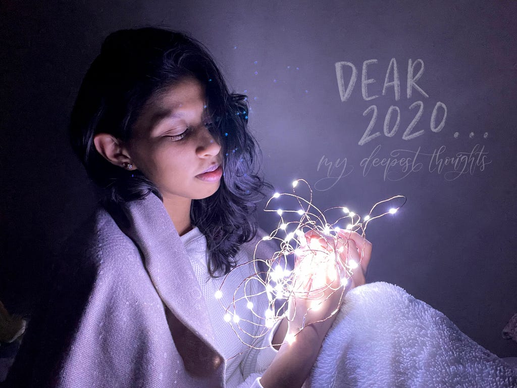Sitting with fairy lights that are in a ball, wearing a scarf. Labeled, “Dear 2020…my deepest thoughts”.