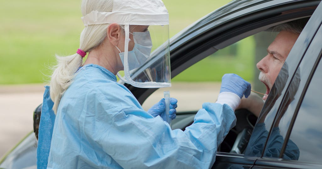 Dressed in full protective gear, a healthcare worker collects a sample from a man sitting inside his car for COVID testing.